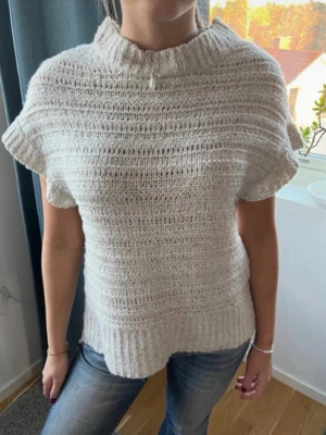 Beige stickad topp med kort ärm - Säljer en beige stickad topp med korta ärmar och hög ribbad krage. Tröjan har ett grovt stickat mönster och bred ribb vid nederkanten. Perfekt att styla med jeans för en avslappnad look.