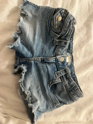 Blå jeansshorts med fransig kant - Säljer ett par blå jeansshorts med fransig, rå kant och dekorativa fickor med knappdetaljer både fram och bak. Klassisk femficksmodell med bälteshällor och slitna detaljer för en avslappnad look. Perfekta för varma dagar! jätte små på mig så dom kommer aldrig till användning längre!