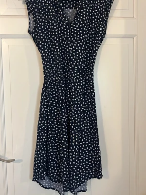 Flörtig sommarklänning - Söt marinblå kortklänning med vitt blommönster och v-ringad hals. Den har en snöre som man kan nyta vid median samt fickor. Klänningen är ärmlös och har en lätt utställd kjol som ger en flowy känsla. Perfekt för varma dagar och enkel att styla med sneakers eller sandaler.