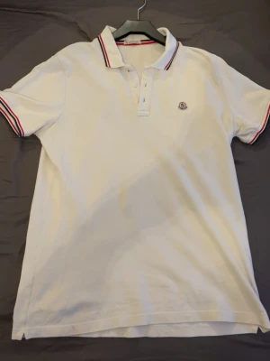 Vit Moncler pikétröja med rödblå detaljer - Klassisk vit pikétröja från Moncler med rödblå ränder på krage och ärmslut. Tröjan har knappslå med tre knappar och Moncler-logga broderad på bröstet. Tillverkad i pikématerial som ger en sportig och stilren look.