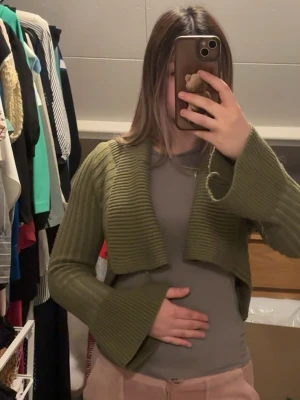 Olivgrön cropped kofta med vida ärmar - Snygg olivgrön cropped kofta i ribbstickat material. Koftan har långa, vida ärmar och en öppen front som ger en trendig look. Perfekt att slänga över en topp för en cool layering-effekt.