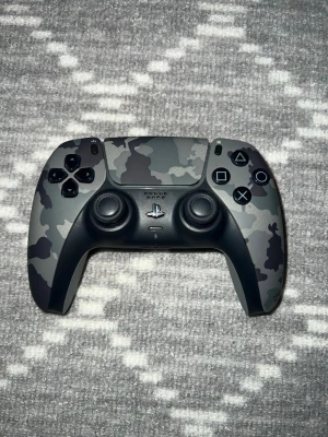 PS5 DualSense Camo handkontroll - Snygg PlayStation 5 DualSense handkontroll i grått camomönster. Ergonomisk design med klassiska PlayStation-knappar, två analoga spakar och touchpad. Tillverkad i slitstark plast och passar perfekt för gaming på PS5.