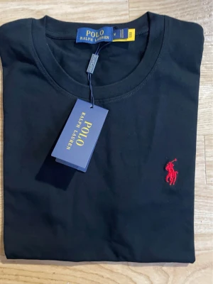 Svart t-shirt från Polo Ralph Lauren - Klassisk svart t-shirt från Polo Ralph Lauren med rund hals och korta ärmar. T-shirten har den ikoniska röda logotypen broderad på bröstet och är tillverkad i mjuk bomull för en skön känsla. Perfekt till jeans eller shorts.