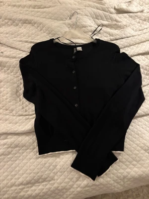 Svart kofta från DIVIDED, S - En svart, klassisk kofta från DIVIDED med rund halsringning och knäppning framtill. Långärmad och i mjukt, tunt material.