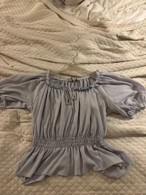 Ljuslila blus från Gina Tricot - Supersöt ljusblå blus från Gina Tricot med resår i midjan och volangkant nedtill. Blusen har puffiga trekvartsärmar, rynkad urringning med knytband och mjukt, tunt tyg som ger en luftig känsla.