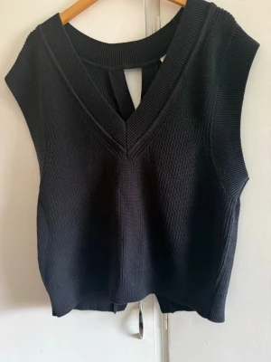 Svart ribbad croppad stickad topp - Snygg svart croppad stickad topp med bred v-ringning och ärmlös design. Toppen har en cool öppen rygg med twistad detalj som ger en unik look. Perfekt att styla med höga jeans eller kjol för en trendig outfit.