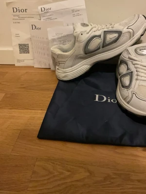 Vita Dior B30 sneakers - Säljer ett par vita Dior B30 sneakers med gråa och silvriga detaljer. Skorna har en chunky sula, meshpaneler och stora CD-loggor på sidorna. Klassisk låg modell med snörning och sportig vibe. Kommer med dustbag och originalhandlingar.