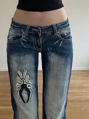 Blue queen extreme low waist W26 jeans - Säljer ett par blå bootcut jeans med slitningar och en stor vit tribal brodyr på vänster lår. Jeansen har låg midja, fem fickor och kontrastsömmar. Materialet är klassisk denim i bomull. Perfekta för dig som gillar en edgy och unik stil.