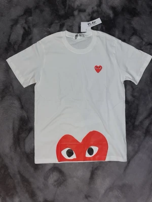 Vit Comme des Garçons Play t-shirt - Vit t-shirt från Comme des Garçons Play med det ikoniska röda hjärtat med ögon tryckt både litet på bröstet och stort nedtill. Helt ny, säljer pga att jag köpte fel storlek. Den är storlek Large men sitter som Medium