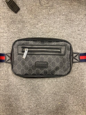 Svart magväska från Gucci - Snygg svart magväska från Gucci med klassiskt GG-mönster, silverfärgad dragkedja och läderdetaljer. Väskan har ett justerbart band i rött och blått och en praktisk ytterficka med dragkedja framtill. Perfekt för att bära det viktigaste med stil.