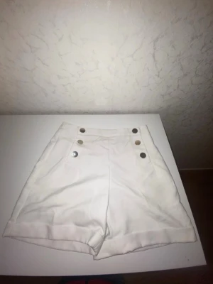 Vita högmidjade shorts med knappar - Stilrena vita shorts med hög midja och dekorativa silverfärgade knappar framtill. Klassisk design med uppvikta benslut och en ficka bak. Perfekta för varma dagar och enkel att matcha med olika toppar.