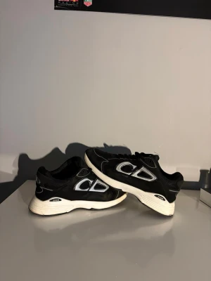 Svarta Dior sneakers med vit sula - Snygga svarta Dior sneakers med vit tjock sula och silverfärgade detaljer på sidan. Skorna har en sportig design med lågt skaft och är tillverkade i en mix av textil och syntetmaterial. Perfekta för dig som vill ha en trendig och bekväm sneaker med exklusiv känsla. Med dust bag