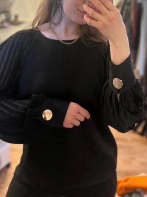 River Island svart blus med plisserade ärmar - Stilren svart blus med vida, långärmade ärmar i transparent tyg och stora guldknappar vid manschetterna. Blusen har rund halsringning och en loose passform som ger en avslappnad men chic look. Perfekt för dig som gillar trendiga detaljer och elegant stil.