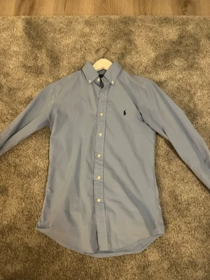 Blå skjorta från Polo Ralph Lauren - Klassisk ljusblå skjorta från Polo Ralph Lauren med button-down krage och broderad logga på bröstet. Skjortan har långa ärmar och vita knappar framtill. Perfekt för en clean och stilren look.