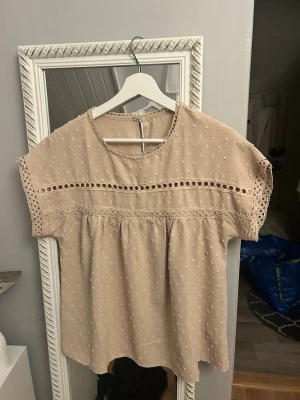 Beige blus med spetsdetaljer - Söt beige blus med korta ärmar och rund halsringning. Blusen har broderade prickar, spetsdetaljer och dekorativa hålband över bröstet och ärmarna. Luftig och avslappnad passform, perfekt till jeans eller kjol.