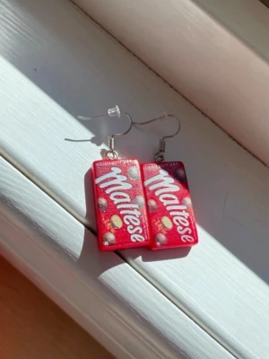 Maltesers örhängen - Unika örhängen formade som små Maltesers-lådor i röd plast med vita och gula detaljer. Rektangulär form och tydlig logga framtill. Perfekta för dig som gillar godis och vill sticka ut med accessoarer som syns.