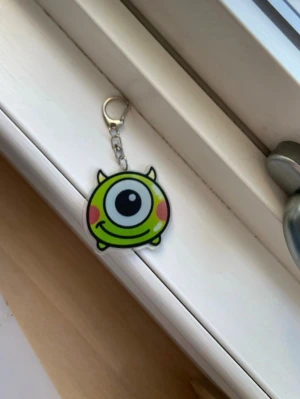 Grön monster nyckelring - Säljer en söt nyckelring med ett grönt monster med ett stort öga och små horn. Nyckelringen har en metallhake och själva figuren är gjord i hård plast. Perfekt att hänga på väskan eller nycklarna för en lekfull touch.