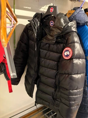 Svart dunjacka från Canada Goose - Snygg svart dunjacka från Canada Goose med klassisk logga på ärmen och dragkedja framtill. Jackan har en quiltad design och huva, perfekt för kalla vinterdagar. Tillverkad i glansigt material som ger en modern look.