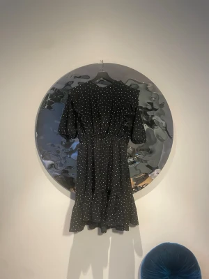 Svart prickig kortklänning med volang - Svart kortklänning med vita prickar och volang nertill. Klänningen har korta puffärmar och en markerad midja med resår. Materialet känns lätt och luftigt, perfekt för dig som gillar en söt och trendig look.