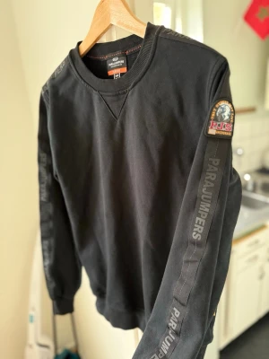 Svart sweatshirt från Parajumpers - Svart sweatshirt från Parajumpers med rund halsringning och ribbade muddar. Tröjan har tryckt logga på ärmen och ett snyggt patch på axeln. Tillverkad i mjuk fleece för extra komfort. Perfekt för en avslappnad och stilren look.