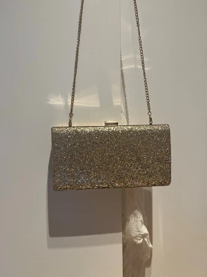 Glittrig guld kuvertväska med kedja - En snygg kuvertväska i rektangulär form med glittrigt guldigt material och metallkedja. Väskan har ett metallspänne upptill och hela ytan är täckt av glitter som ger ett lyxigt intryck. Perfekt för att lyfta din outfit.