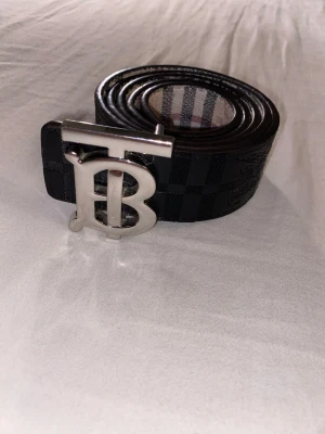 Svart Burberry bälte med silverdetaljer - Snyggt svart bälte från Burberry med klassiskt rutmönster och präglad riddarlogga. Bältet har ett stort, silverfärgat TB-spänne som ger en lyxig känsla. Perfekt accessoar för att lyfta din outfit.