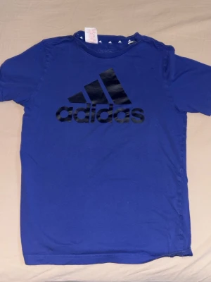 Blå Adidas t-shirt med logga - Säljer en blå t-shirt från Adidas med stor svart logga på bröstet. T-shirten har klassisk rund hals och korta ärmar, perfekt för dig som gillar sportiga och stilrena plagg. Materialet är mjukt och skönt bomull som passar till många olika outfits.