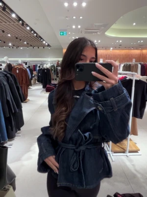 Mörkblå jeansjacka med bälte - Snygg mörkblå jeansjacka med markerad midja tack vare det breda knytbältet. Jackan har vida ärmar och klassiska sömmar i kontrastfärg. Perfekt för dig som vill ha en trendig och avslappnad look.