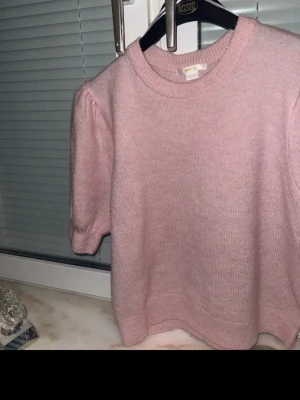 Ljusrosa stickad puffärmströja - Söt ljusrosa stickad tröja med puffärmar och rund halsringning. Tröjan har en croppad passform och ribbade muddar vid ärmslut och nederkant. Perfekt för dig som gillar en mjuk och trendig look.