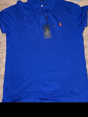 Blå pikétröja från Polo Ralph Lauren - Klassisk blå pikétröja från Polo Ralph Lauren med korta ärmar och orange broderad logga på bröstet. Tröjan har krage, knappar vid halsen och är tillverkad i mjuk bomull. Perfekt till en avslappnad men snygg stil.