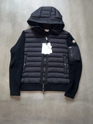 Svart pufferjacka med huva från Moncler - Snygg svart pufferjacka från Moncler med vadderad front och huva. Jackan har stickade ärmar, ribbade muddar och en ficka med dragkedja på ena ärmen. Stängs med dubbel dragkedja framtill och har Moncler-logga på ärmen. Perfekt för kalla vinterdagar. Storleken är L men sitter som m eftersom moncler är små i storlekarna.