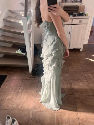 Ljusgrön långklänning med volanger - Superfin ljusgrön långklänning med tunna axelband och volangdetaljer längs hela klänningen. Klänningen har en draperad urringning och är tillverkad i ett lätt, flowy material. Bra i längden för mig som är 170 cm. Den går ända ner till golvet men släpar inte för mycket i marken. Endast använd 1 gång. Har gjort en liten ”söm” på insidan av klänning vid bysten för att den skulle sitta bättre. Skulle tro att den enkelt kan sprättas upp. Nypris 1070 kr. Bara att skicka meddelande om du undrar nått. 