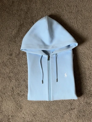 Ljusblå hoodie från Polo Ralph Lauren - Snygg ljusblå hoodie med dragkedja från Polo Ralph Lauren. Endast använd 1-2 gånger. Tvättas innan den skickas, sällsynt, storlek S. Pris kan diskuteras vid snabbaffär mvh 47 😃