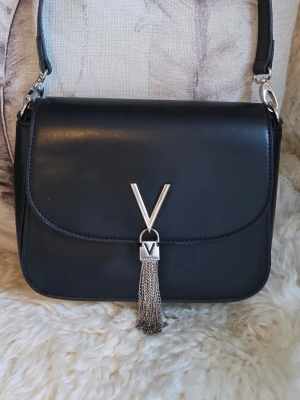 Valentino - Stilren svart handväska från Valentino i slätt läder med silverfärgad V-detalj och dekorativa kedjor framtill. Perfekt accessoar för dig som gillar klassisk men trendig design. Den är i fint skick 