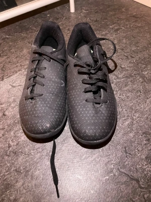 Svarta futsal/gympaskor - Säljer ett par svarta sneakers med låg profil och klassisk snörning. Skorna har en diskret mönstrad mesh-överdel som ger bra andningsförmåga och en tunn, flexibel sula. Perfekta för dig som gillar stilrena och bekväma skor till träning eller vardagsaktiviteter. Skorna saknar sula!!