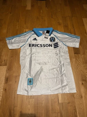 Marseille home kit 98/99 - Snygg vit Marseille matchtröja från Adidas med ljusblå detaljer och svart Ericsson-tryck på bröstet. Klassisk krage i blått och klubbmärke på vänster bröst. Tillverkad i lätt och ventilerande material, perfekt för fotboll eller träning.