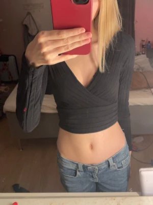 Svart omlottopp med lång ärm - Snygg svart croppad omlottopp med långa ärmar och ribbad struktur. Djup v-ringning och figurnära passform som framhäver midjan. Perfekt till jeans eller kjol för en trendig look.