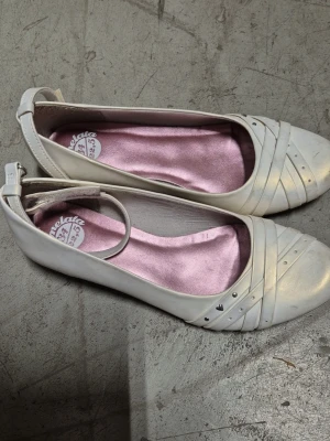 Ballerinaskor - Snygga beige ballerinaskor från Marmelata med dekorativa remmar och små silverfärgade nitar framtill. Skorna har en rundad tå och en justerbar rem runt hälen. Insidan är fodrad med ljusrosa tyg för extra komfort. Finns fläck.