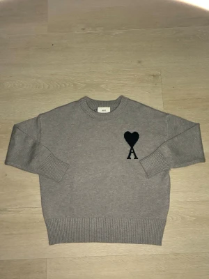 Grå stickad tröja från Ami Paris - Säljer en grå stickad tröja från Ami Paris med svart hjärta och A-logga på bröstet. Tröjan har rund halsringning, ribbade muddar och nederkant. Perfekt för dig som gillar stilrena och trendiga plagg.