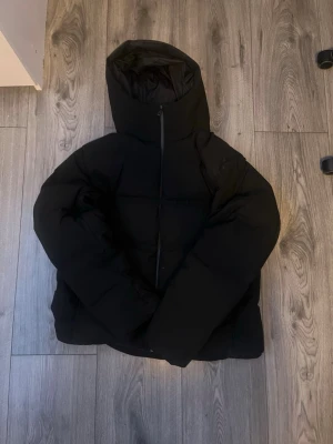 Uniqlo jacka  - Säljer en svart pufferjacka med stor huva och dragkedja framtill. Jackan har en boxig passform och breda kanaler som ger en riktigt puffig look. Perfekt för kalla dagar när du vill hålla dig varm och samtidigt se stilren ut.