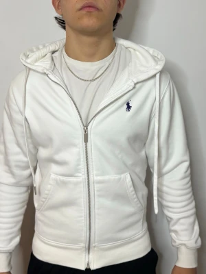 Vit hoodie från Ralph Lauren - Snygg vit hoodie med dragkedja från Ralph Lauren. Klassisk design med blå logga på bröstet, huva med dragsko och två stora fickor framtill. Tillverkad i mjukt material som känns skönt mot huden.