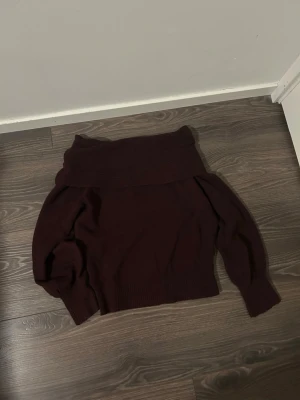 Vinröd off shoulder tröja från NewYorker - Mysig mörkbrun stickad tröja från NewYorker med off shoulder och långa ärmar. Tröjan har ribbstickade detaljer vid kragen, ärmslut och nederkant. Perfekt för att hålla sig varm under kyliga dagar. Tröjan är i storlek 38.
