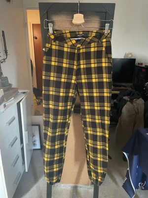 Gula och svarta rutiga byxor - Säljer ett par byxor från American Bazi med svart och gul rutig design. Byxorna har resår vid bensluten och knappstängning i midjan. Perfekta för dig som vill sticka ut med ett coolt mönster och bekväm passform.
