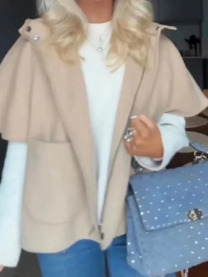 Beige cape-rock med huva - Stilren beige cape-rock med huva och knappar vid axlarna. Rocken har en lös passform och stora fickor framtill. Perfekt för lager-på-lager och ger en trendig siluett. Materialet ser ut att vara mjukt och ull-liknande.