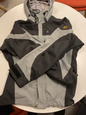 Grå och svart vindjacka från Haglöfs - Snygg vindjacka från Haglöfs i grått och svart med justerbar huva och kardborre vid ärmslut. Jackan har dragkedja framtill, logga på bröstet och coola kontrastpaneler. Perfekt för dig som vill ha en stilren och funktionell jacka till vardags. Pris kan diskuteras 