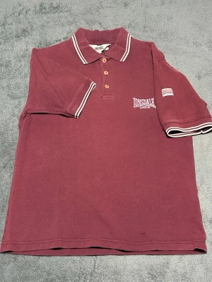 Lonsdale London Polo Shirt – Burgundy – Size S - Klassisk piké från Lonsdale i vinröd färg med vita kontrastdetaljer.  Sportig modell med broderad logga och patch på ärmen, inspirerad av klassisk brittisk street- och sportstil.  Perfekt till jeans eller shorts för en casual look.  Detaljer • Lonsdale London • Burgundy / vinröd färg • Broderad logga • Patch på ärmen • Kortärmad  📦 Skickas snabbt 💬 Hör av dig vid frågor