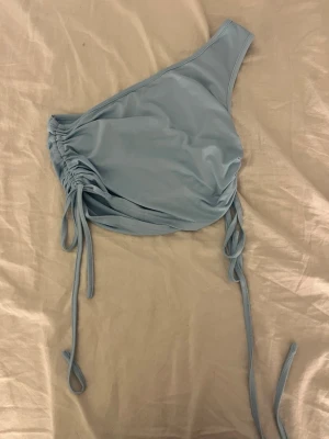 Ljusblå oneshoulder topp med snörning - Trendig ljusblå oneshoulder topp med snygga snörningsdetaljer på sidan. Toppen är croppad och har en tight passform. Tillverkad i mjukt syntetmaterial som ger en skön känsla. Perfekt för dig som vill sticka ut med en unik och modern look.