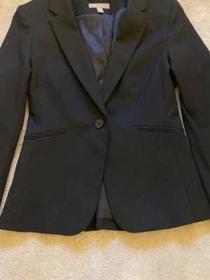 Svart klassisk kavaj med knapp - Stilren svart kavaj med en knapp framtill och två fickor. Kavajen har klassisk krage och är helfodrad för en snygg siluett. Perfekt till skjorta eller t-shirt för en clean look. Materialet ser ut att vara en blandning av polyester och viskos.