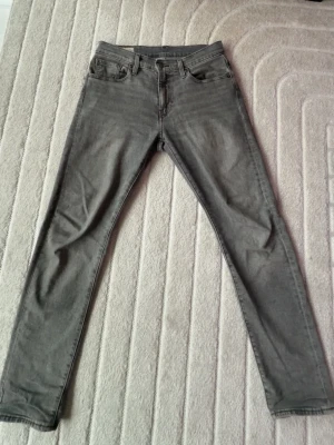 Grå jeans från Levi's - Säljer ett par grå jeans från Levi's med klassisk femficksdesign och raka ben. Jeansen har en snygg tvättad look, läderpatch bak och den ikoniska röda Levi's-taggen på bakfickan. Perfekt för dig som gillar stilrena och tidlösa jeans. modell 502 regular taper