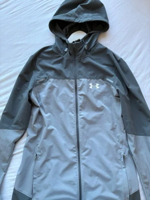  Under Armour Jacka - Snygg grå vindjacka från Under Armour med huva och hel dragkedja framtill. Jackan har två sidofickor med dragkedja och logga på bröstet. Tillverkad i ett lätt och vindtätt material, perfekt för blåsiga dagar. Storlek S men sitter som S/M